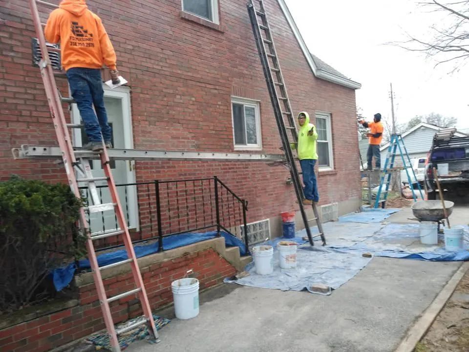 Workers on ladders painting a brick building exterior beside a sidewalk.