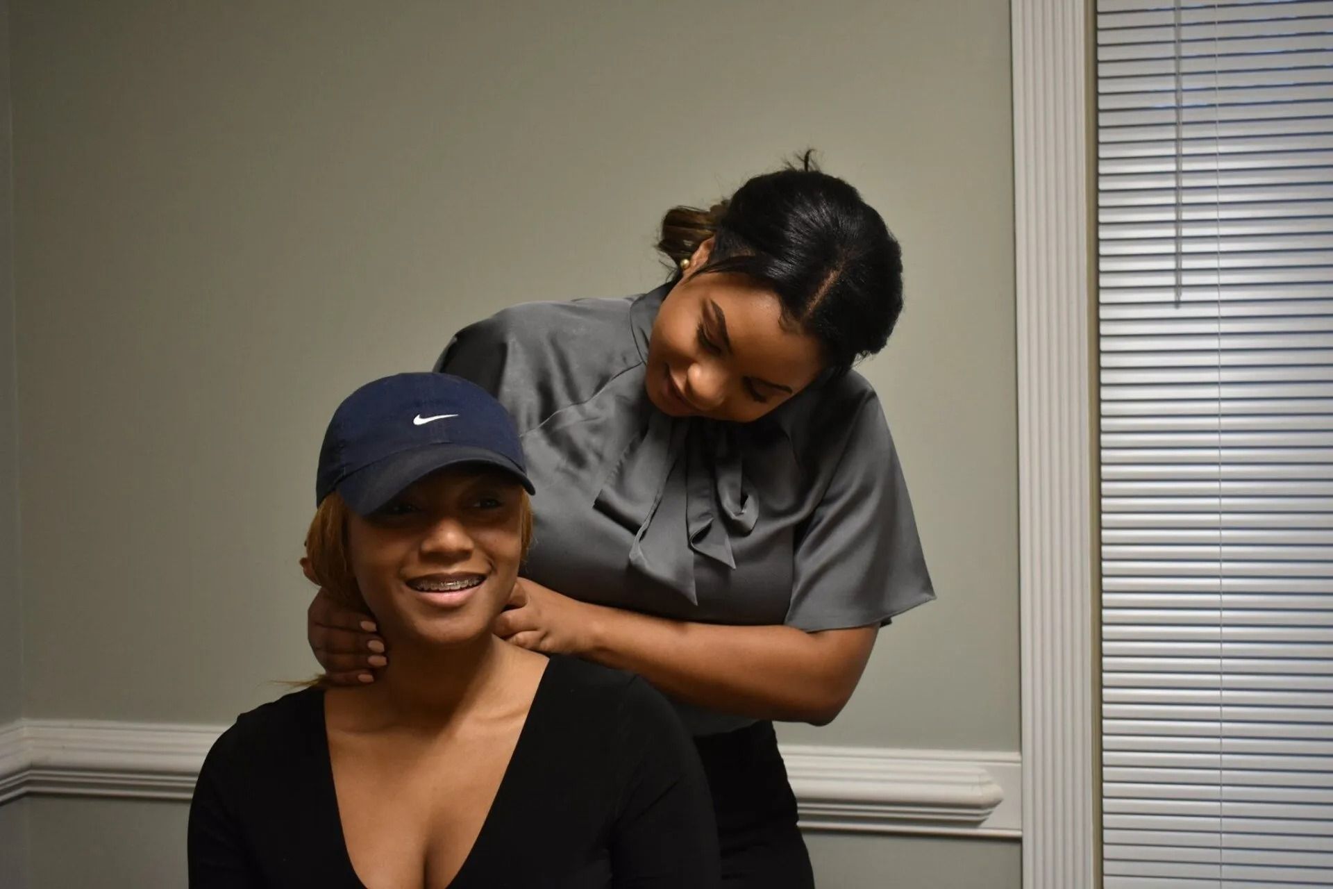 Smiling person sits while another styles their hair in a room with blinds.