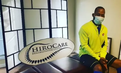 Person in yellow jacket and face mask seated beside HIROCARE sign indoors.