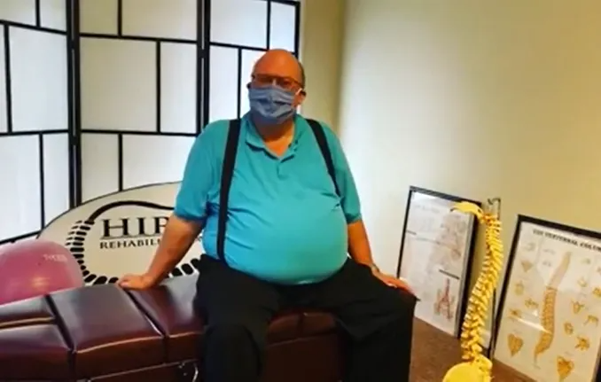 Man seated in a teal shirt and blue mask on a bench beside awards and a sign in an office setting