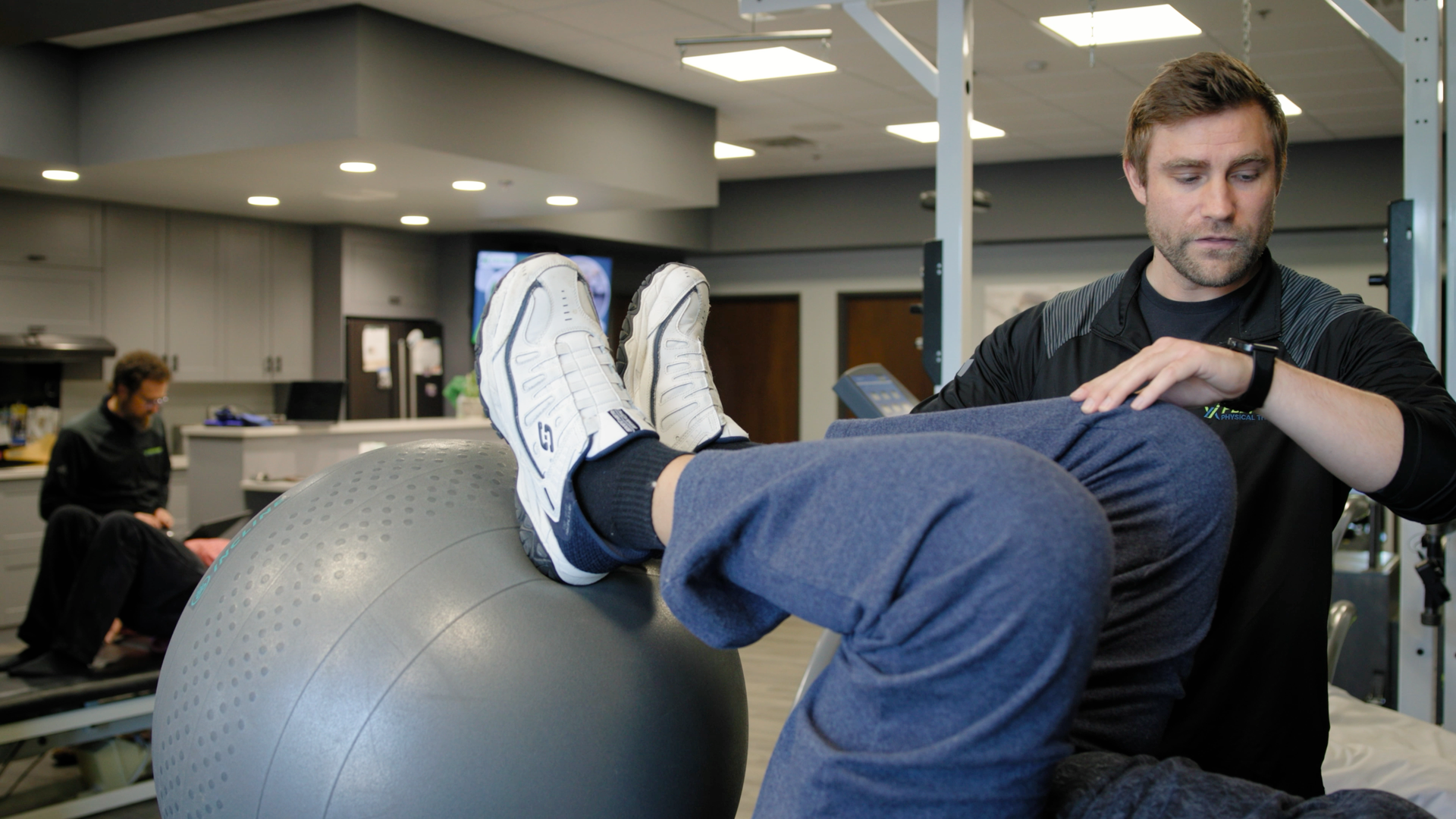 A man is helping another man with a exercise ball in a gym.