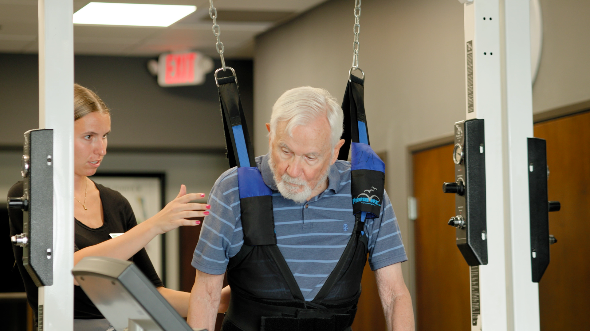 An elderly man is being helped to walk by a woman in a gym.