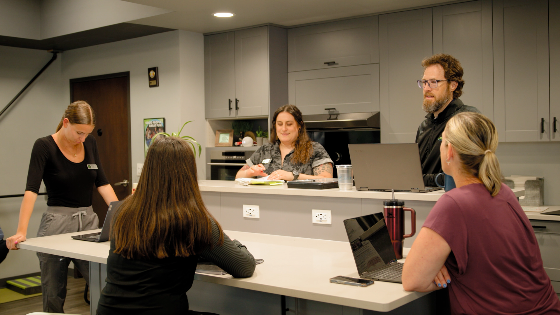 A group of people are sitting at a table in a kitchen with laptops.