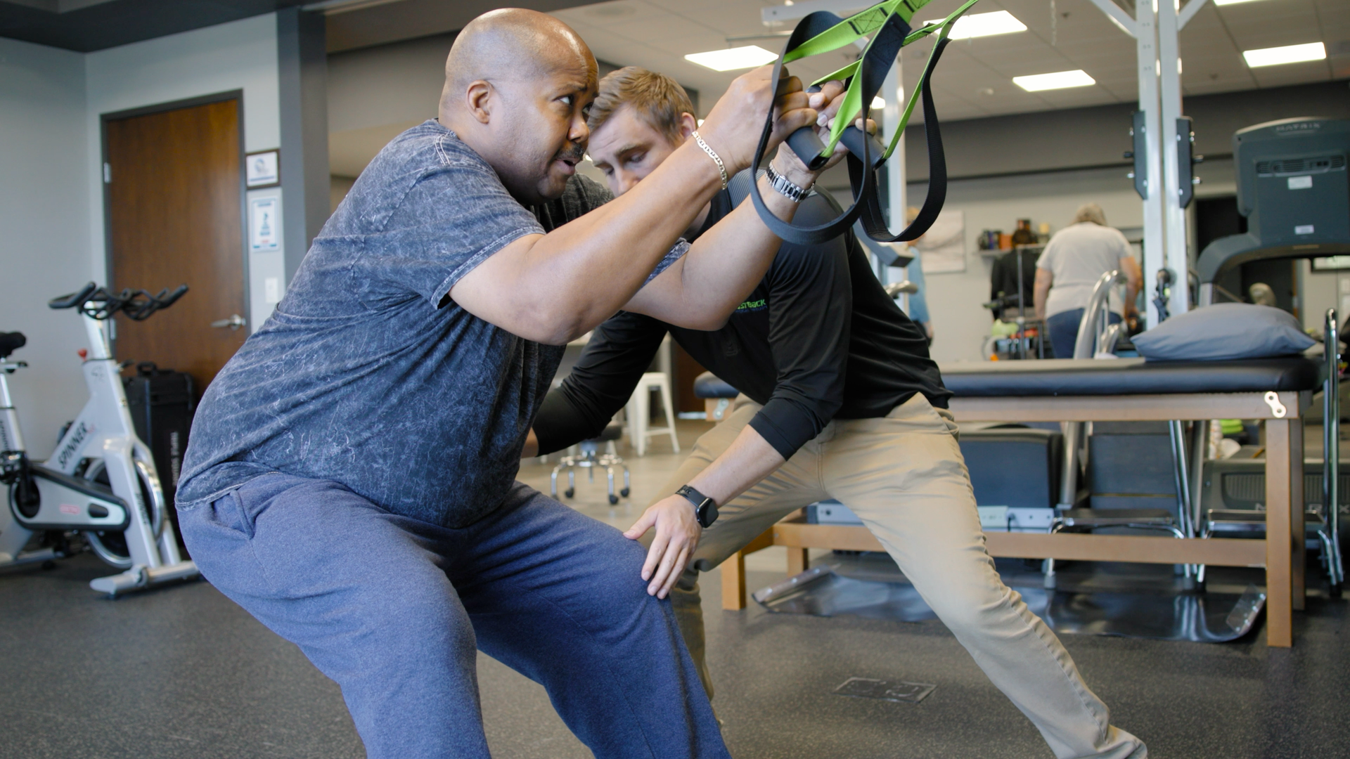 A man is helping another man do squats in a gym.