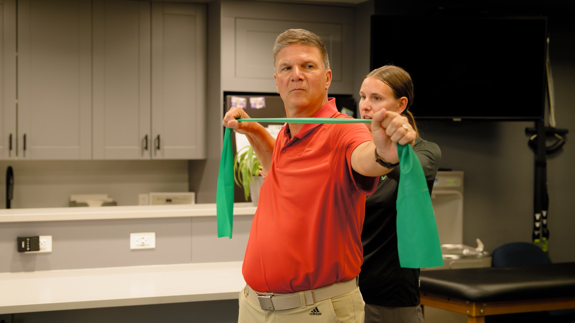 A man in a red shirt is holding a green resistance band over his shoulder.