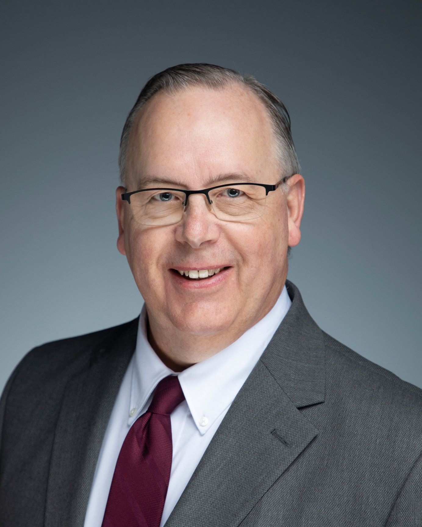 Professional headshot of a smiling man in glasses, gray suit, white shirt, and burgundy tie on a gray background