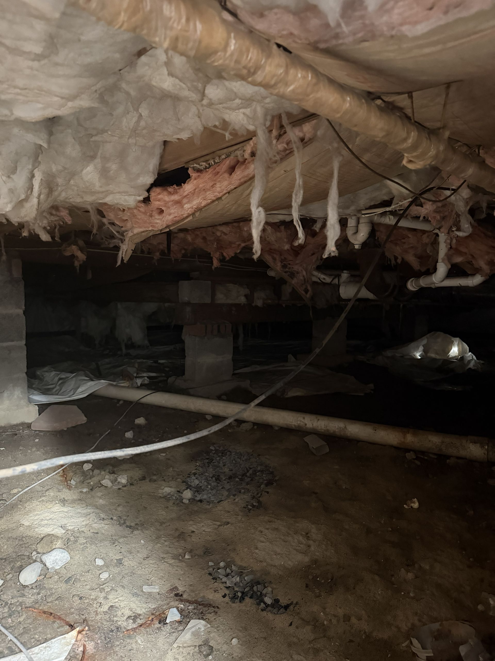 Crawl space with insulation, pipes, and wet ground. Dark, dusty environment with a flashlight.