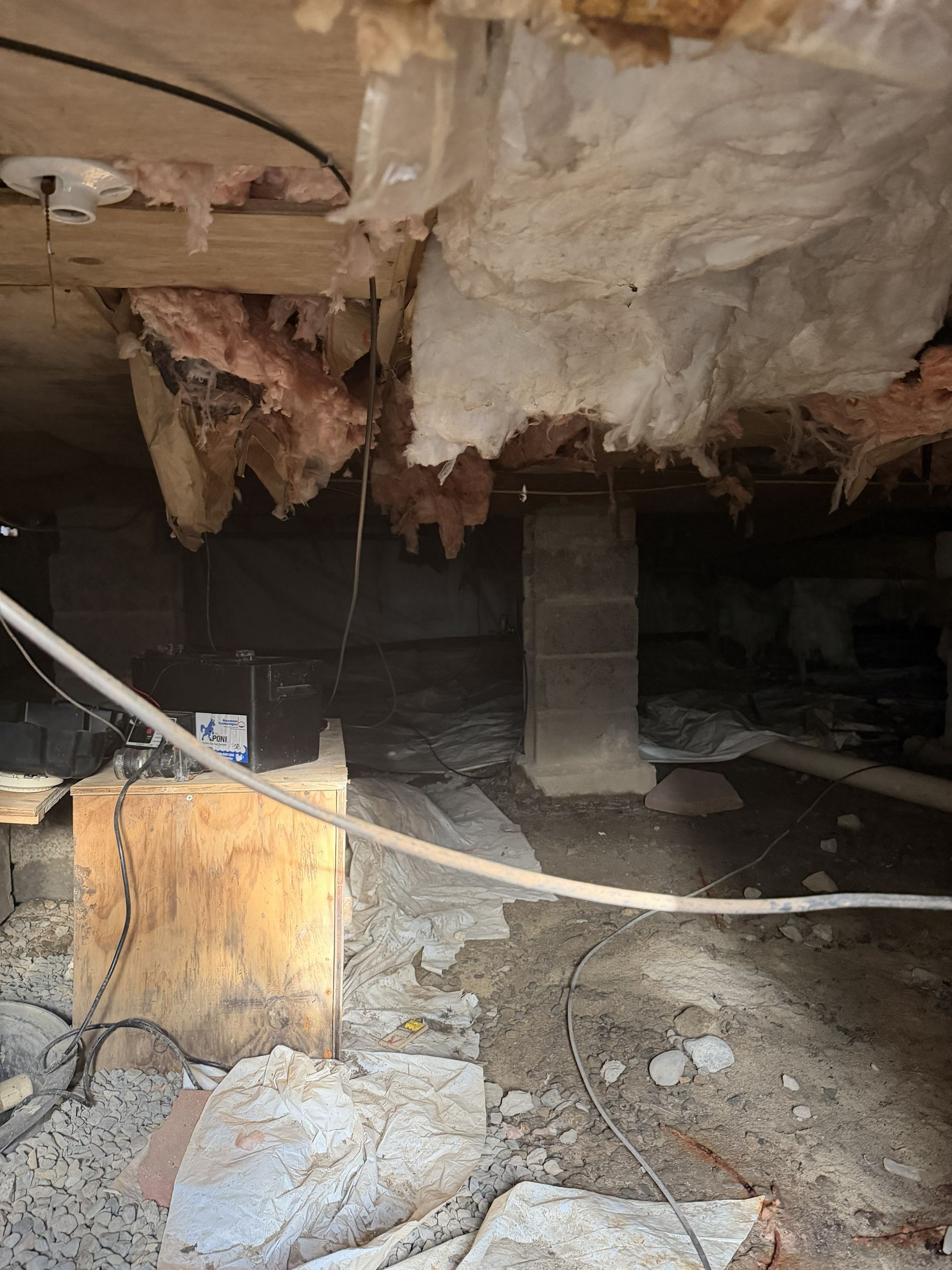 Underneath a house: damaged insulation, support beams, concrete blocks, and debris.