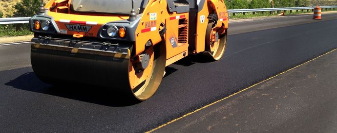 Orange road roller compacting fresh black asphalt on a highway shoulder