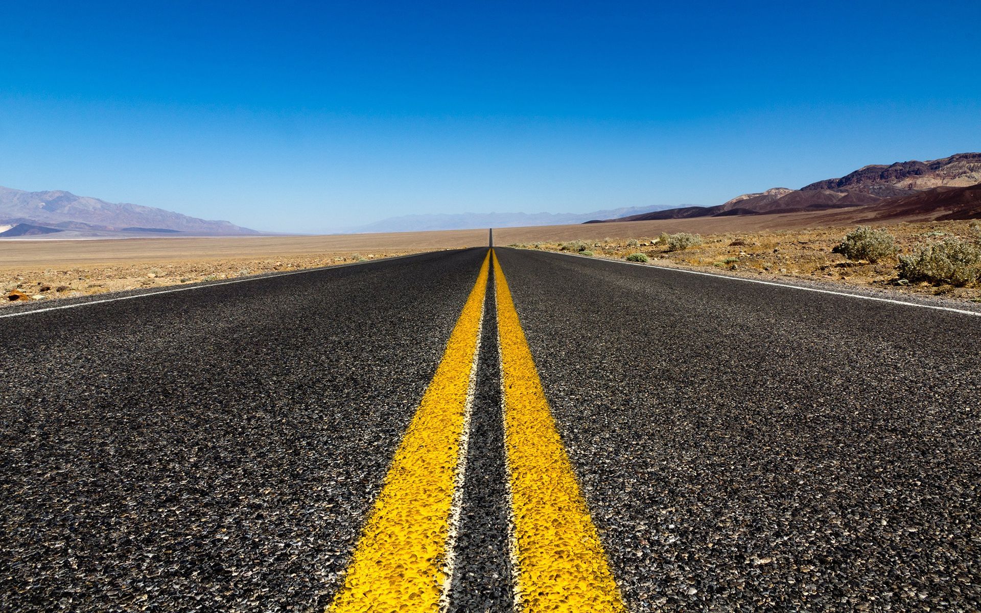 Empty two-lane highway stretching into a desert landscape under a clear blue sky