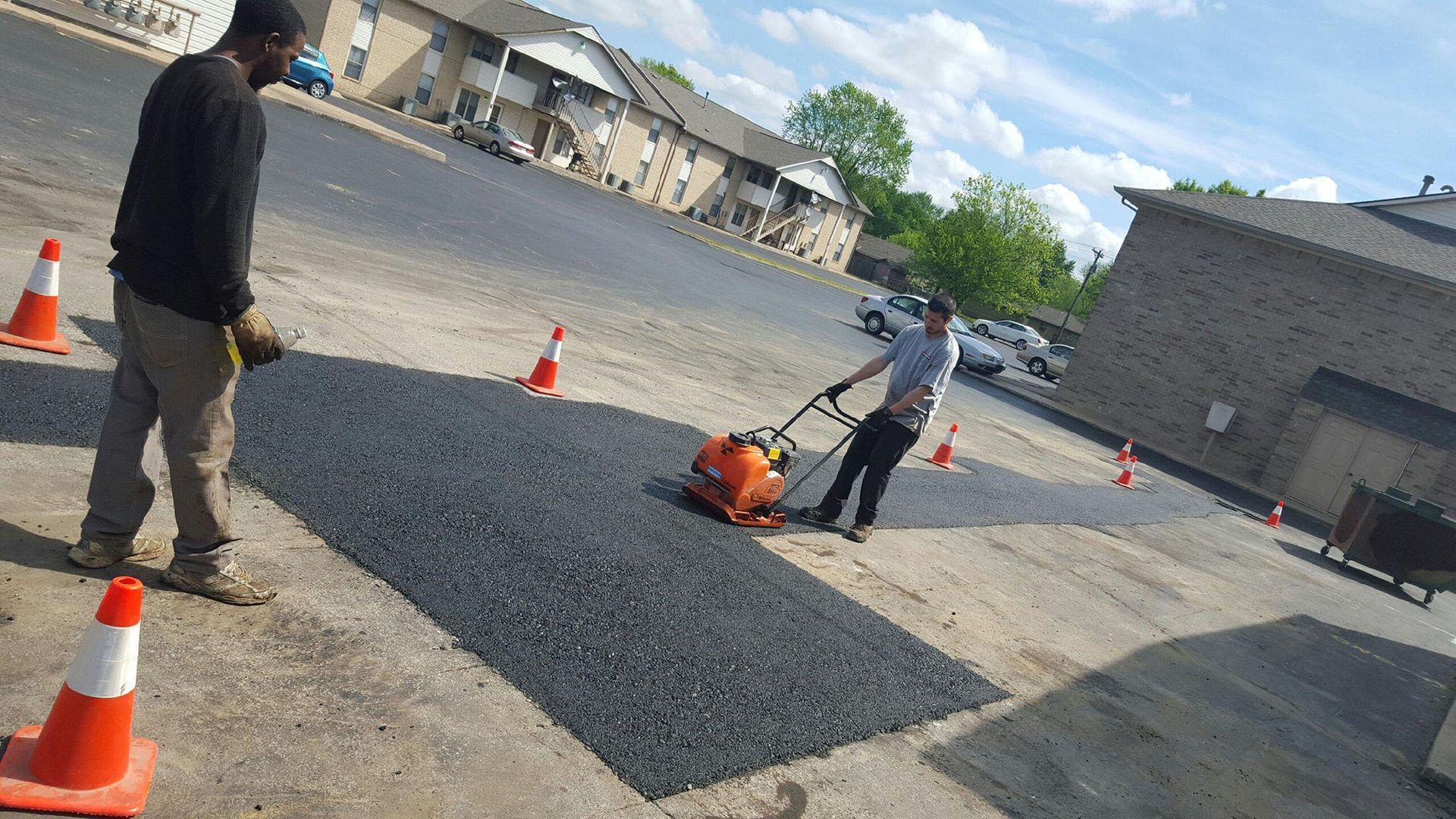Workers paving a gravel lot with orange cones and a compacting machine near a building