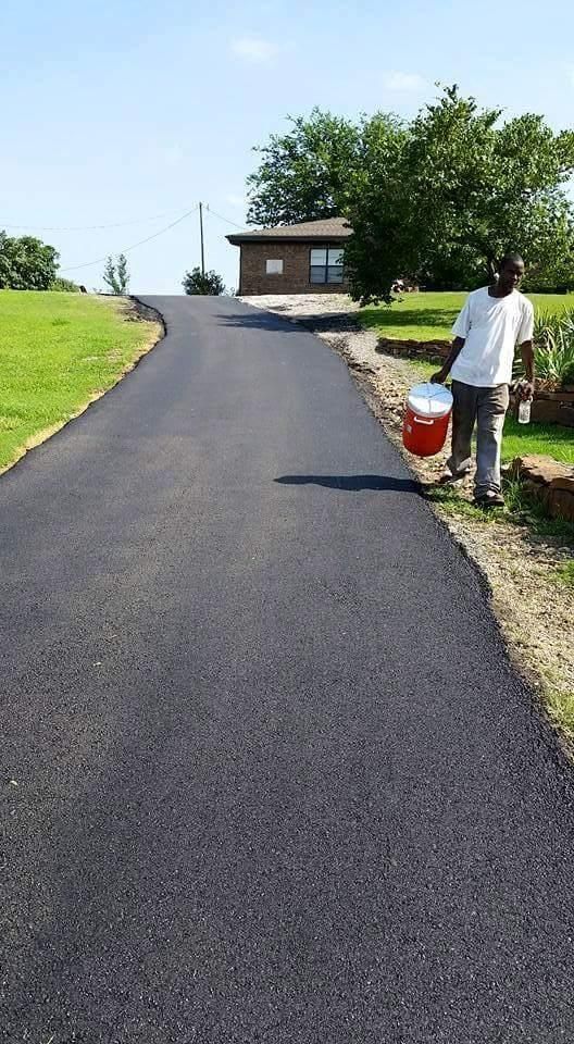 Freshly paved black driveway leading to a small house, with a worker holding a red bucket on the right side
