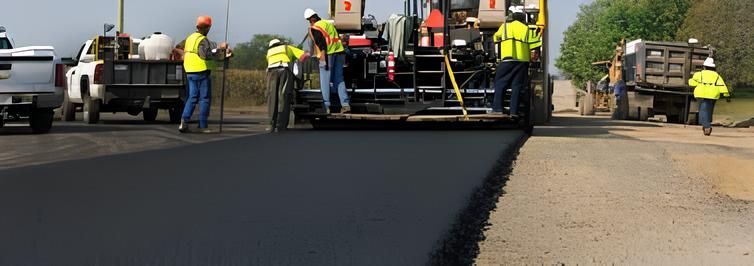 Road crew paving a street, with workers in safety vests beside asphalt machinery and trucks.
