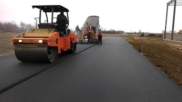 Orange road roller paving a new black roadway beside workers and a dump truck on a rural road