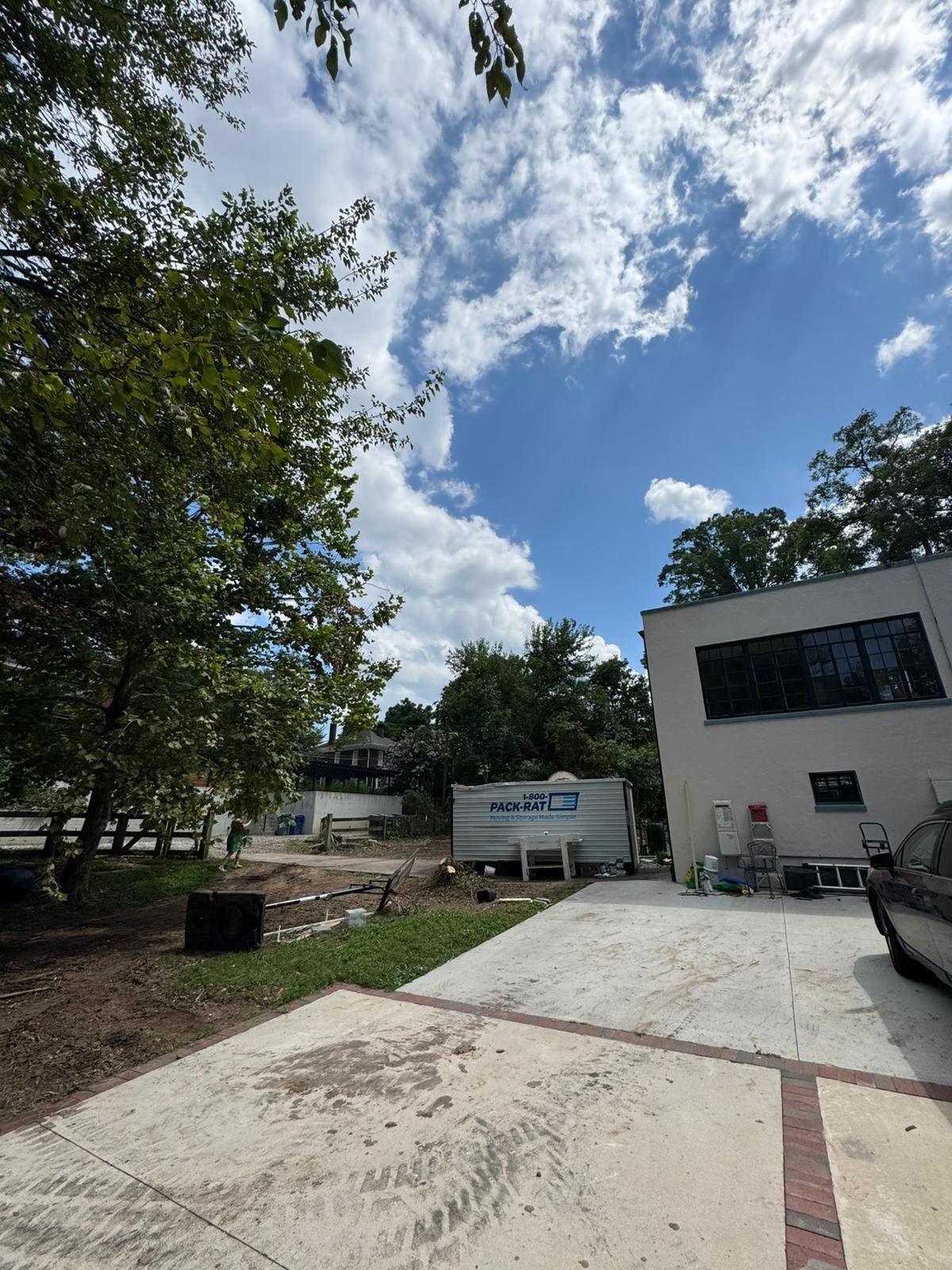 Bright sky with scattered clouds over a building and trees. A concrete driveway is in the foreground. - After