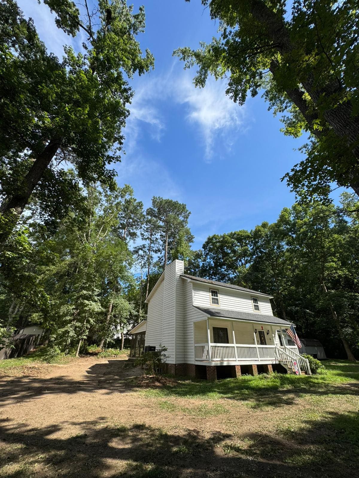 White house with a porch and tall trees against a cloudy sky. The house is on a slight hill. - After