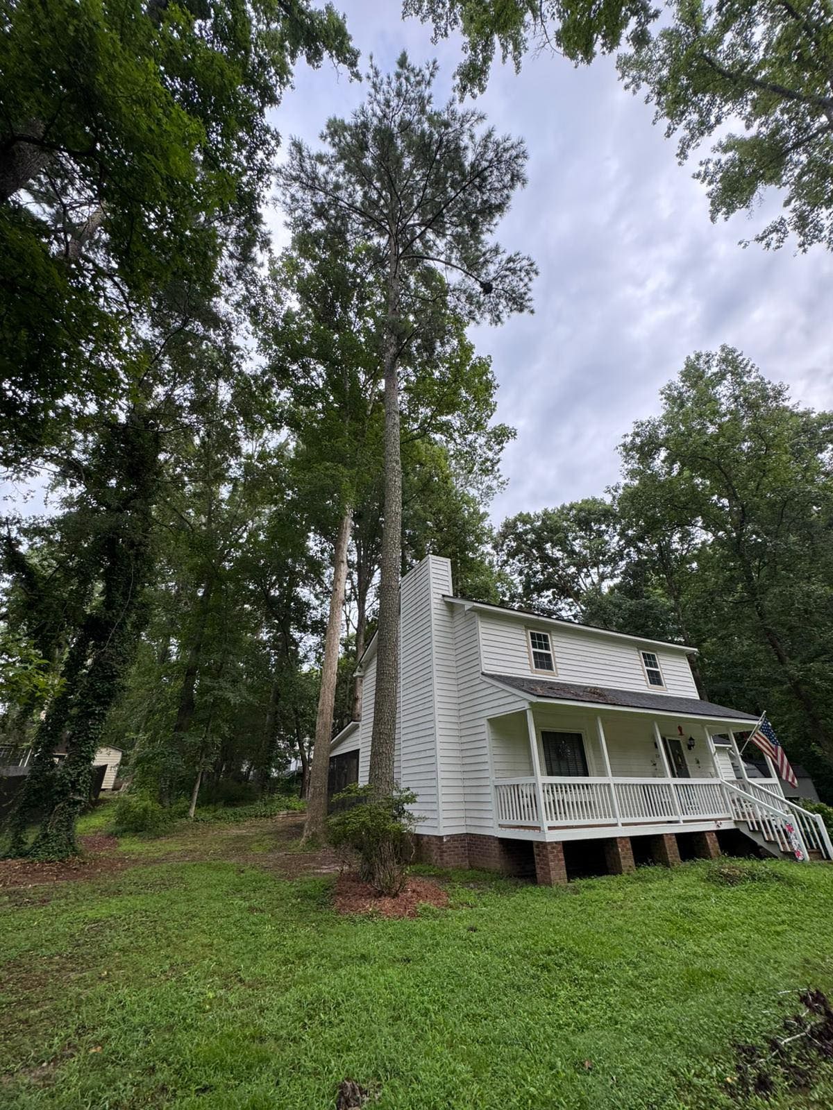 White house with a porch and tall trees against a cloudy sky. The house is on a slight hill. - Before