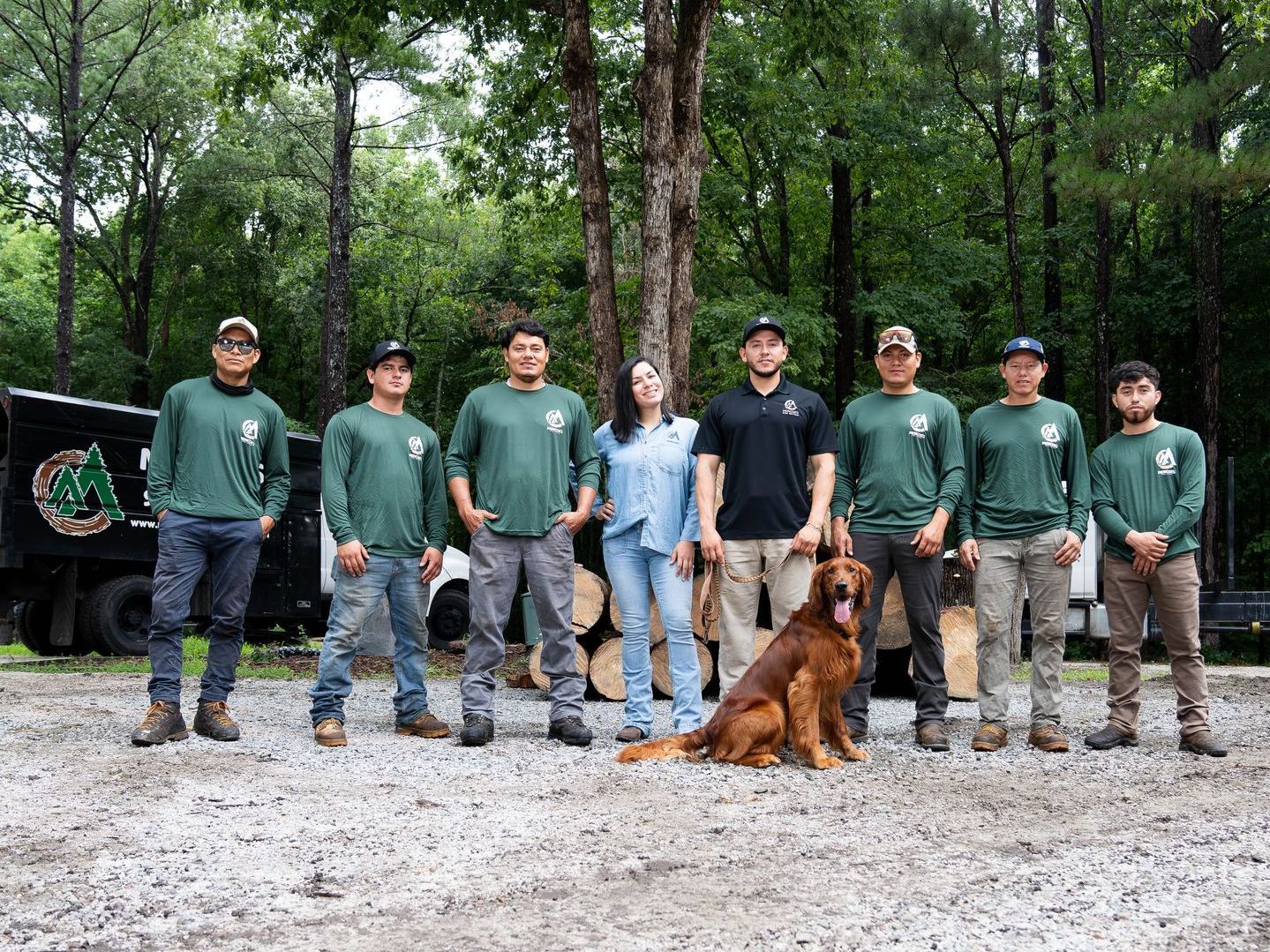 A group of arborists and a dog pose in front of a truck and felled logs in a wooded area.