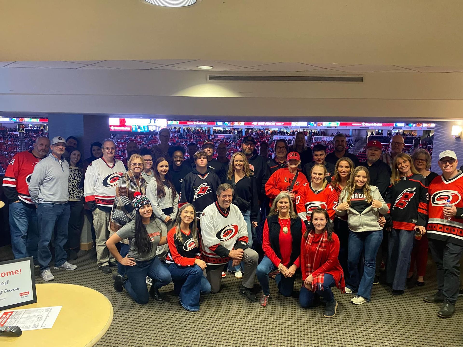 A group of Carolina Hurricanes hockey fans pose together in a viewing area, many wearing team jerseys and cheering.