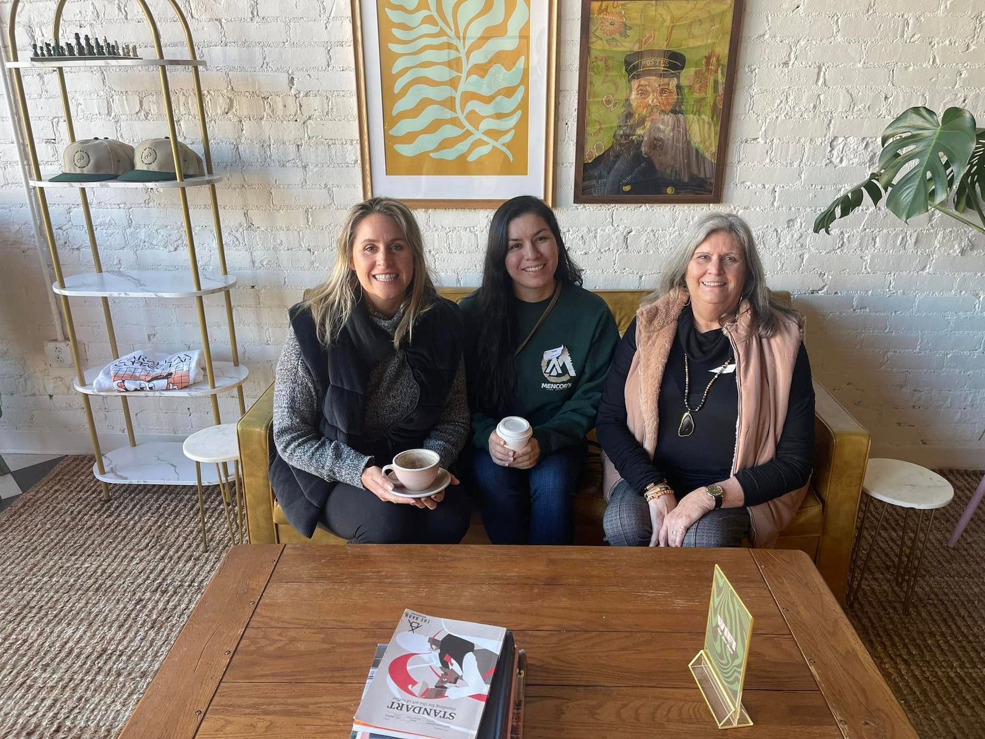 Three women sit on a gold couch in a cafe. One holds a coffee cup, another wears a Harry Potter hoodie. Artwork hung on wall.