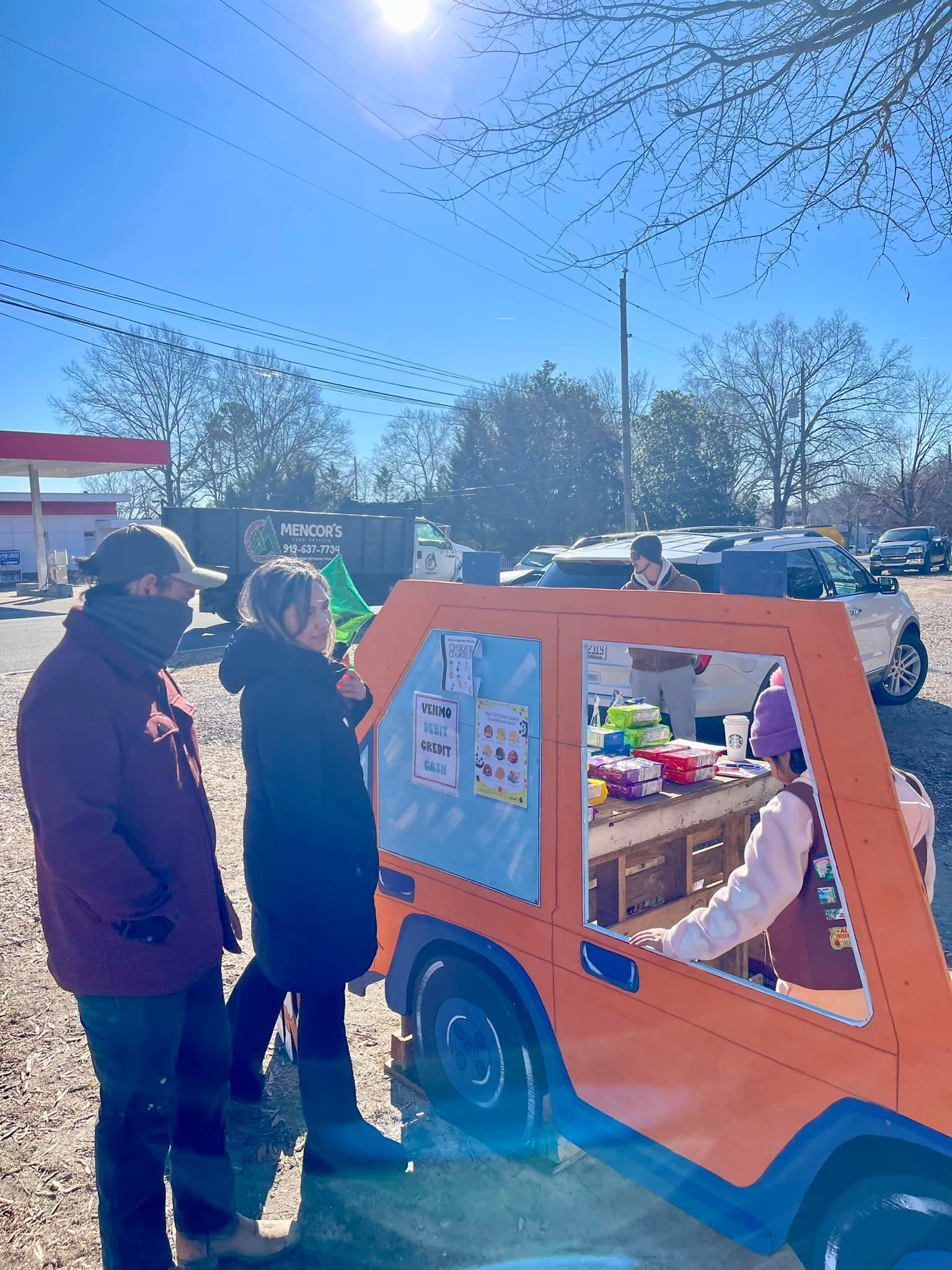 People at a colorful, orange car-shaped stand on a sunny day. One person is selling items, while others look on.