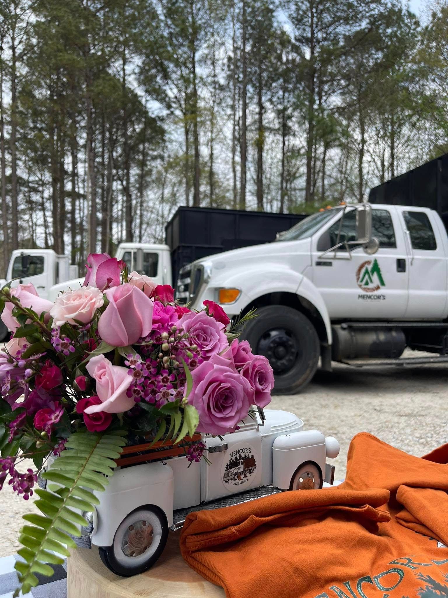 Close-up of a floral arrangement in a toy truck, with white trucks and trees in the background. Roses are pink and purple.