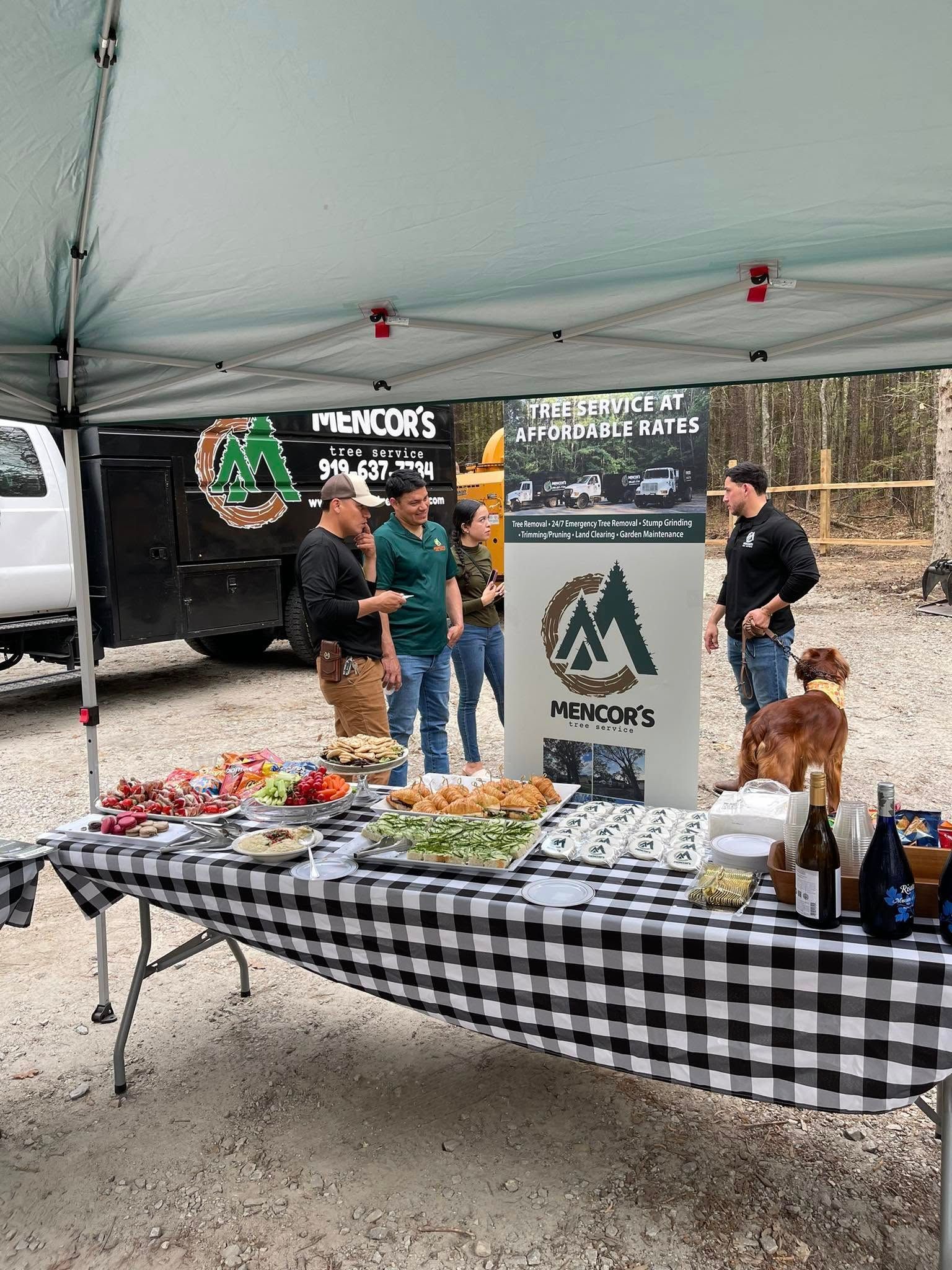A food display under tent with people nearby. The table has a checkered tablecloth, with a company banner in the background.