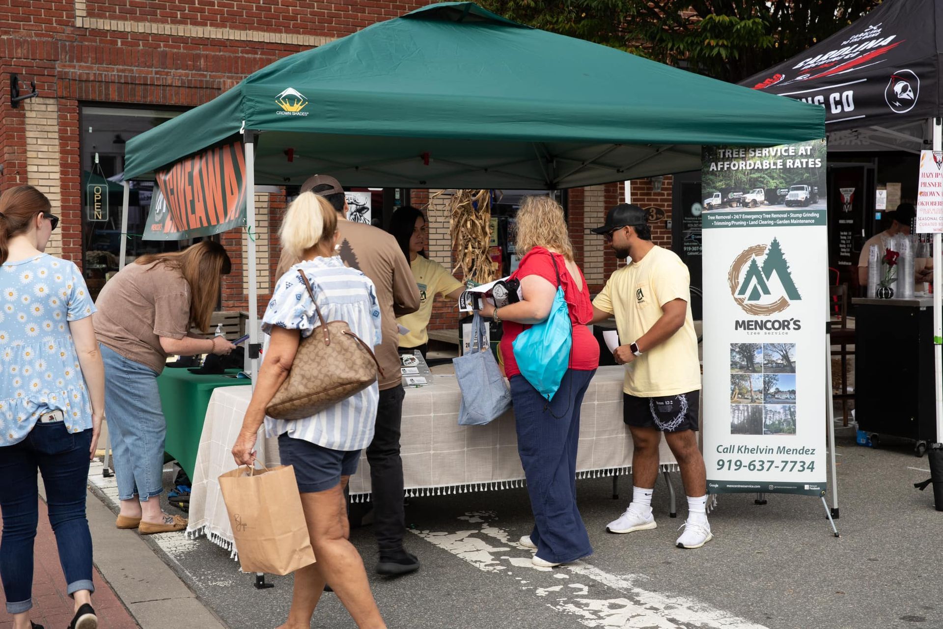 People browse a booth at an outdoor market under a green tent. Several vendors interact with customers.