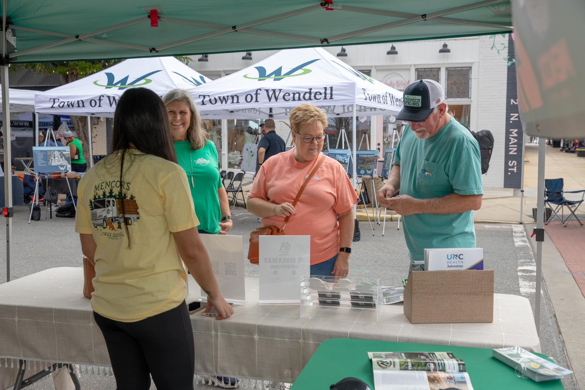 People interacting at a Town of Wendell booth under a tent. A man and woman talk at the table; others stand nearby.