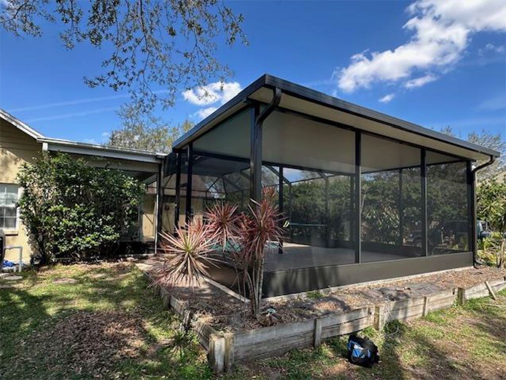Screened-in patio attached to a house, black framing with tan roof, surrounded by vegetation and a raised garden bed.