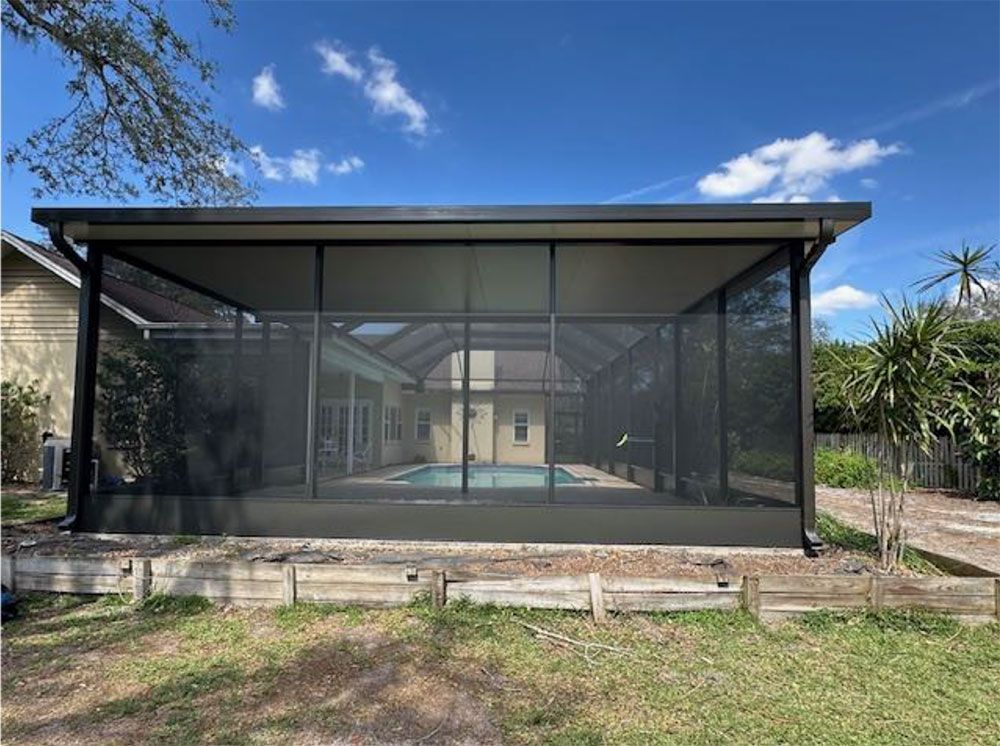Black screened enclosure over a pool, framed by a house and greenery, under a blue sky.