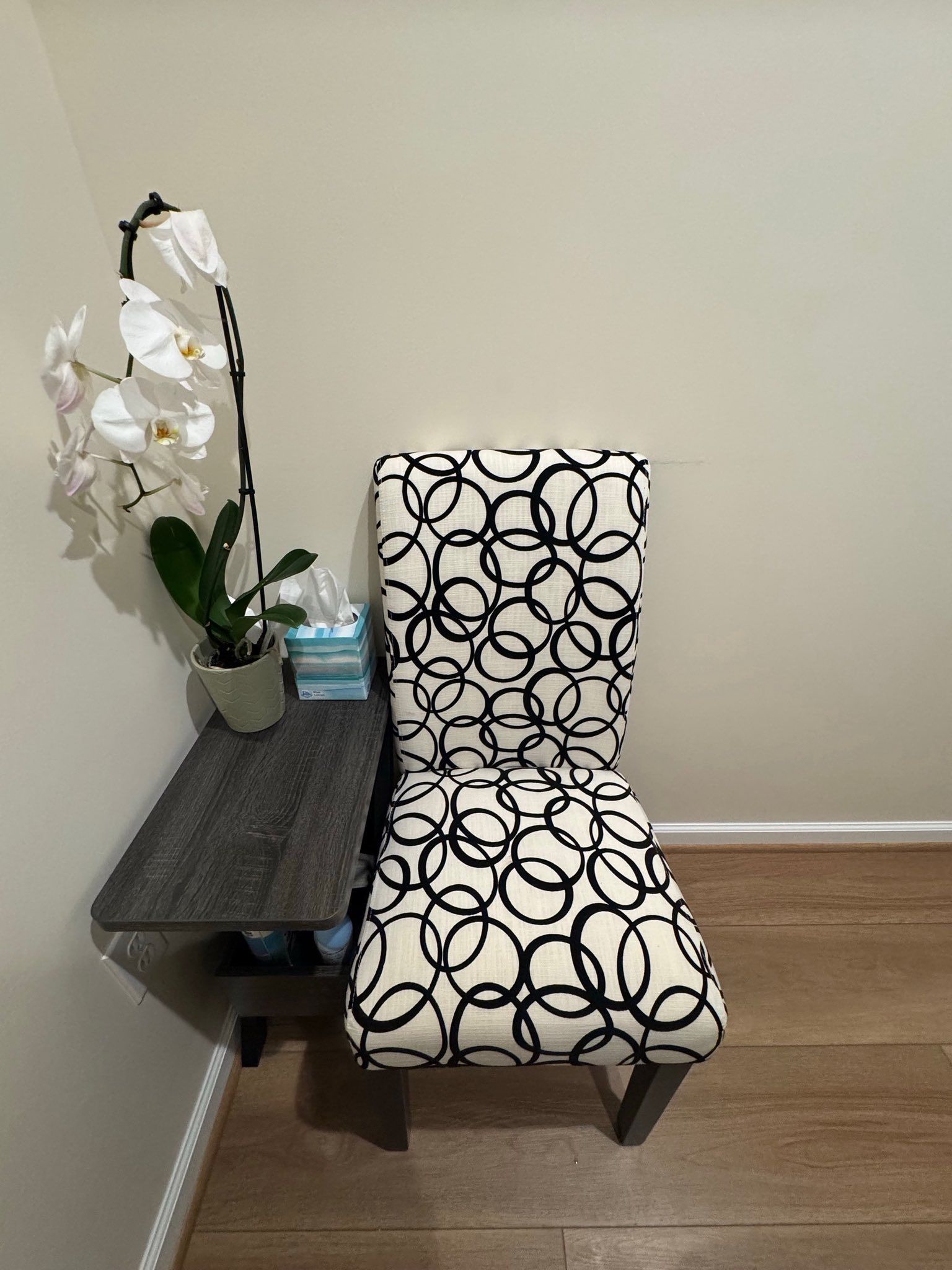 Chair with black and white circles, beside a small table with an orchid, set against a neutral wall.