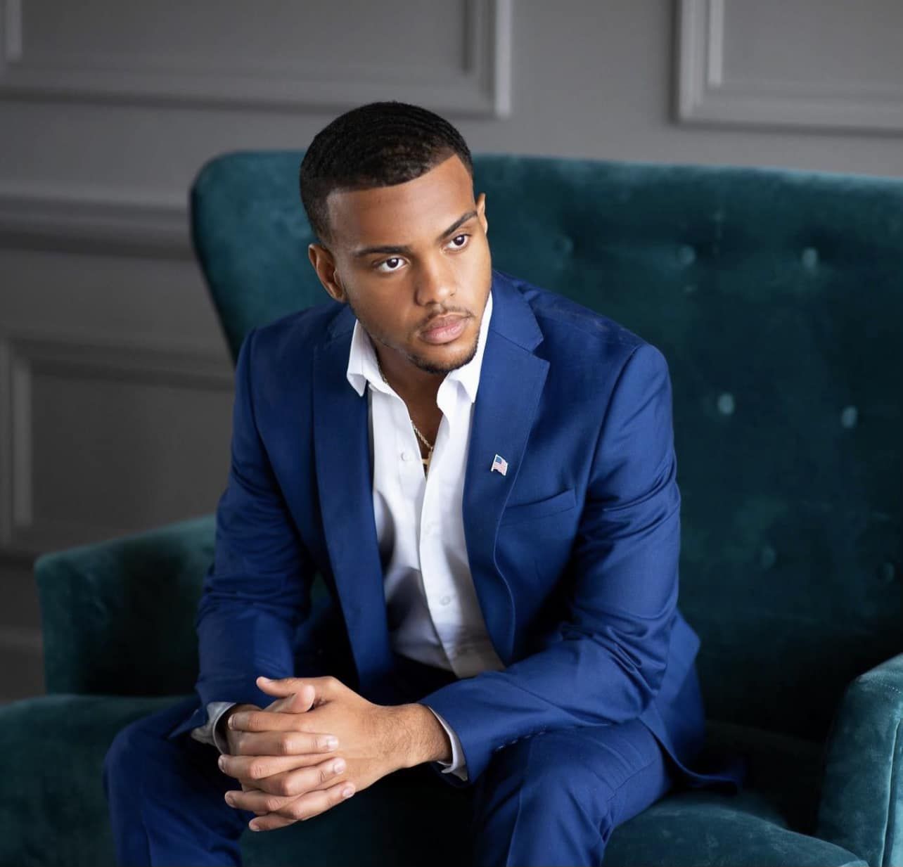 A man in a blue suit is sitting in a chair with his hands folded