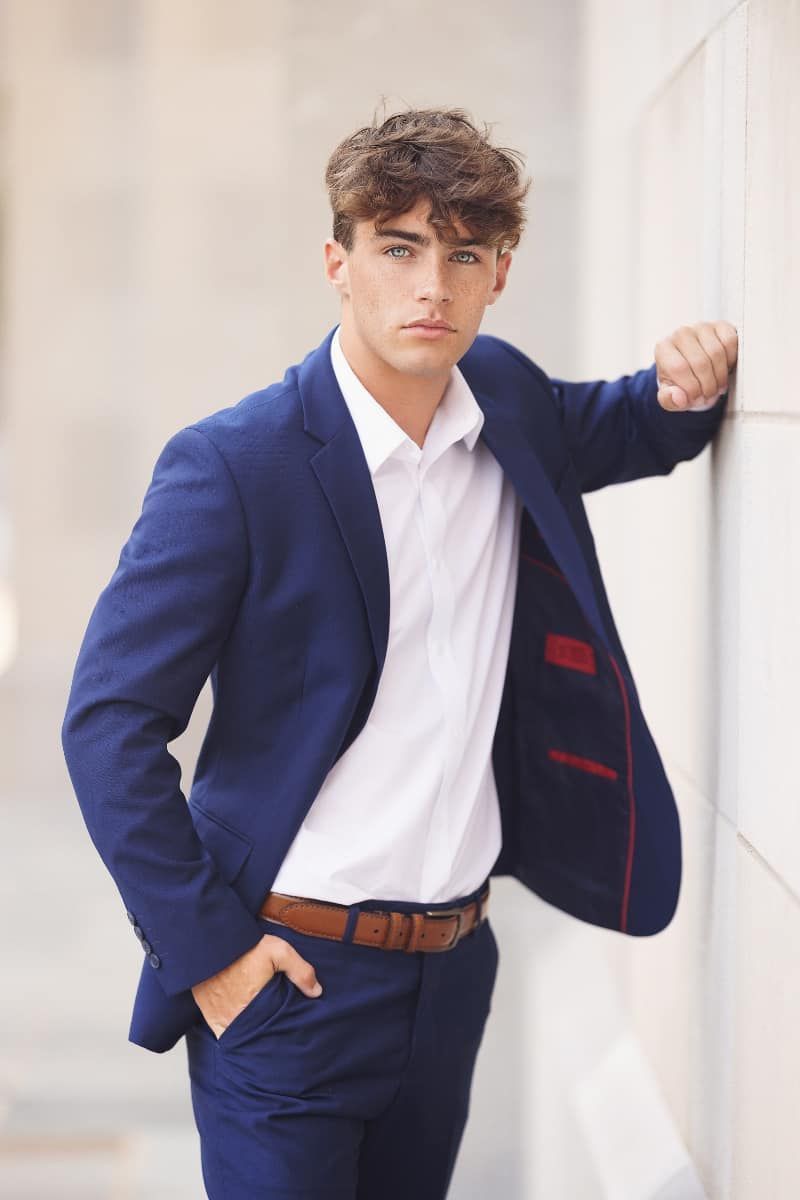 A young man in a blue suit leans against a wall