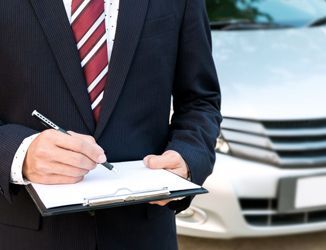 A man in a suit and tie is writing on a clipboard in front of a car