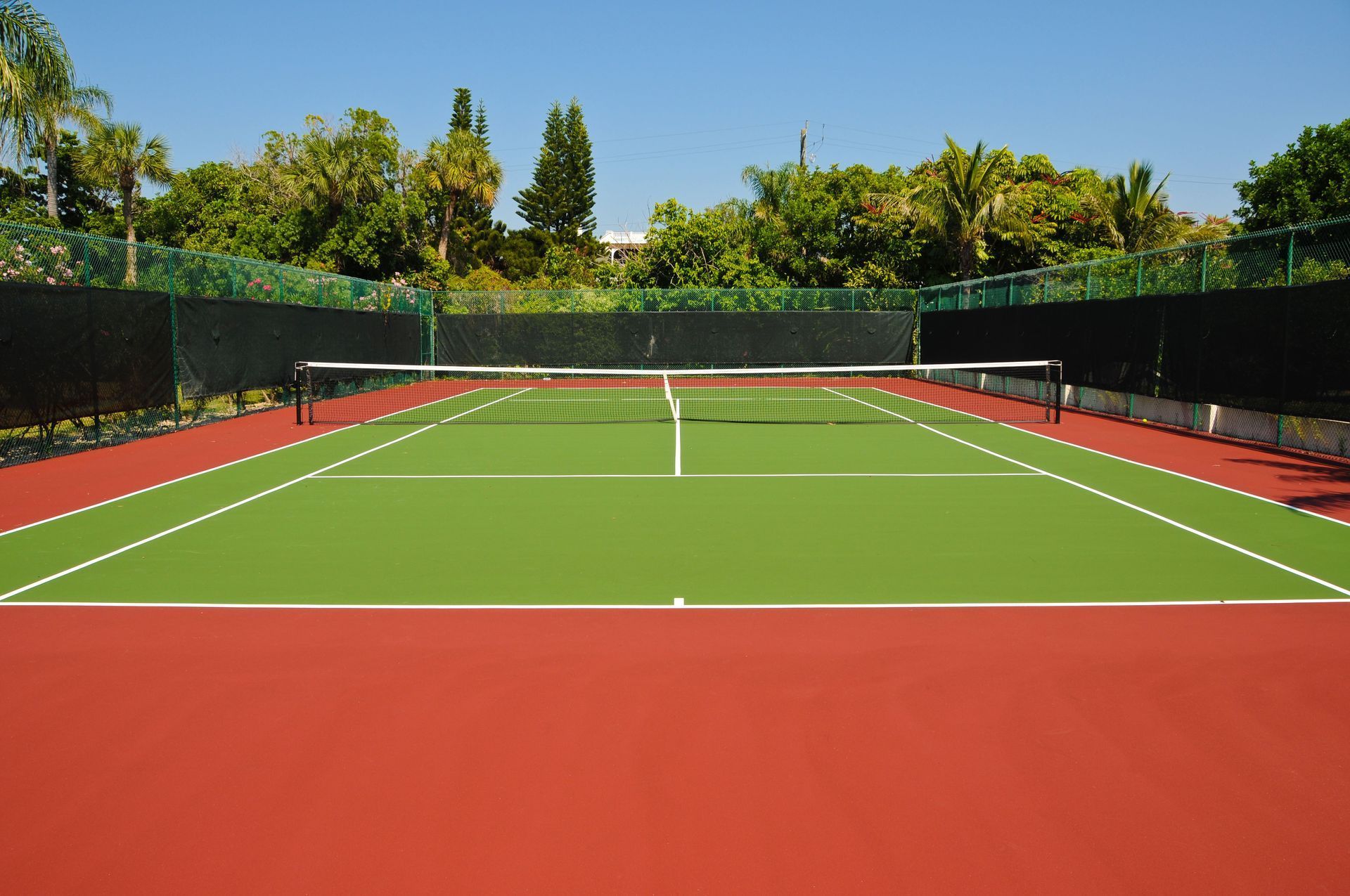 Outdoor tennis court with green playing surface, red borders, net, and trees in the background