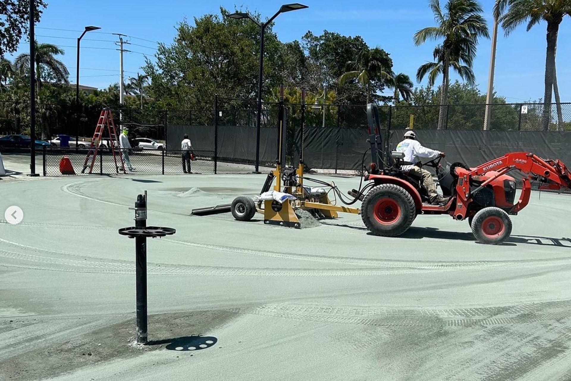 Ground crew operating a red tractor on a tennis court, smoothing the surface under a sunny sky.
