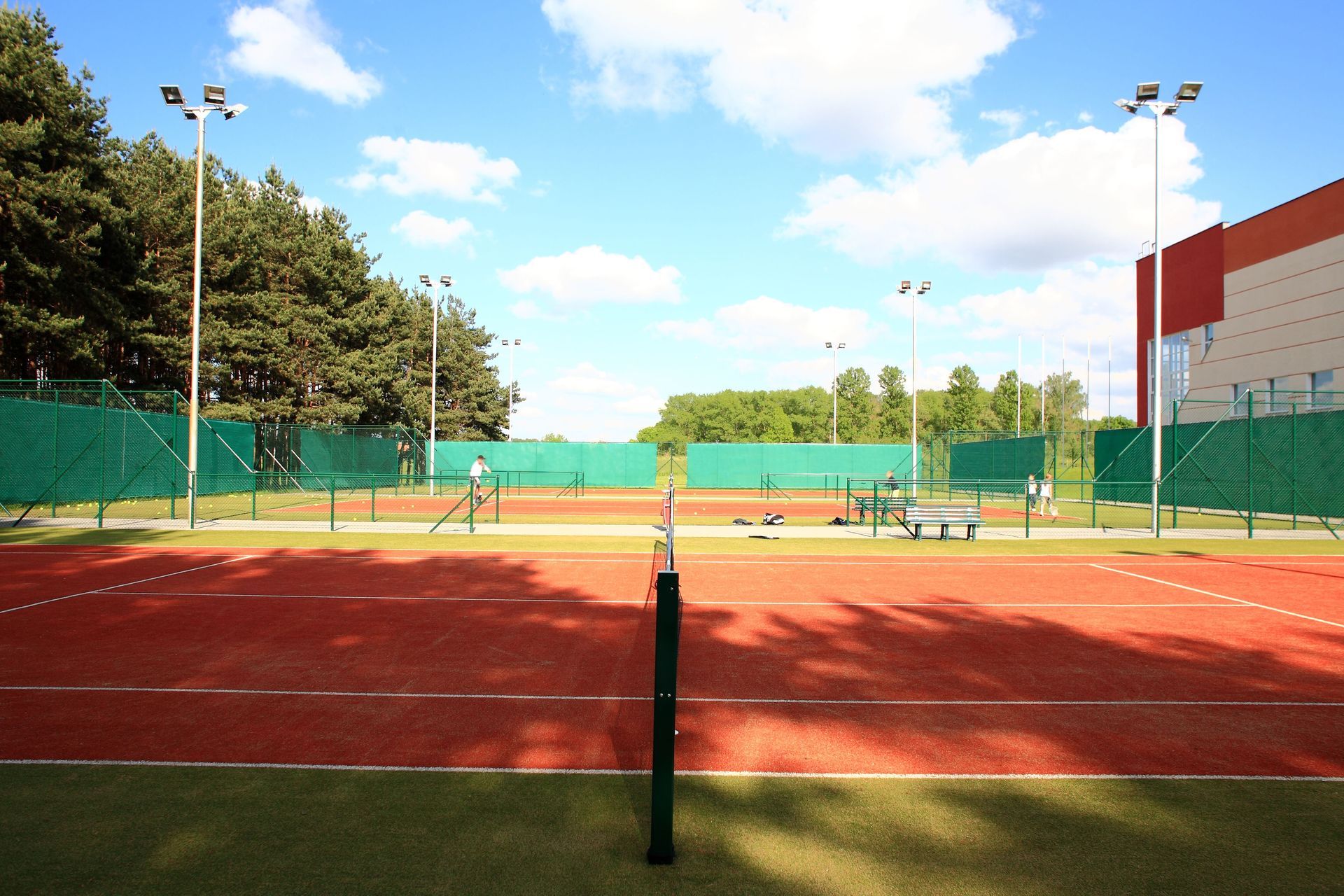 Tennis courts with red clay and green fencing under a blue sky, with a few players in the distance