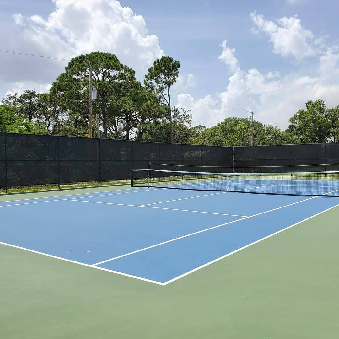 Empty blue tennis court with a net, black fence, and trees under a cloudy sky