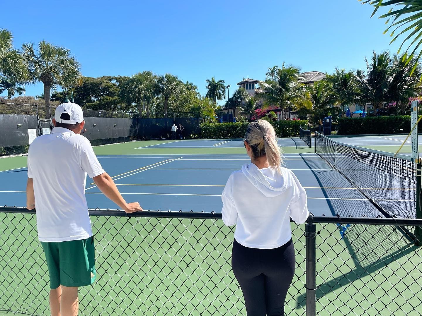 Two people standing by a fenced tennis court under a clear blue sky