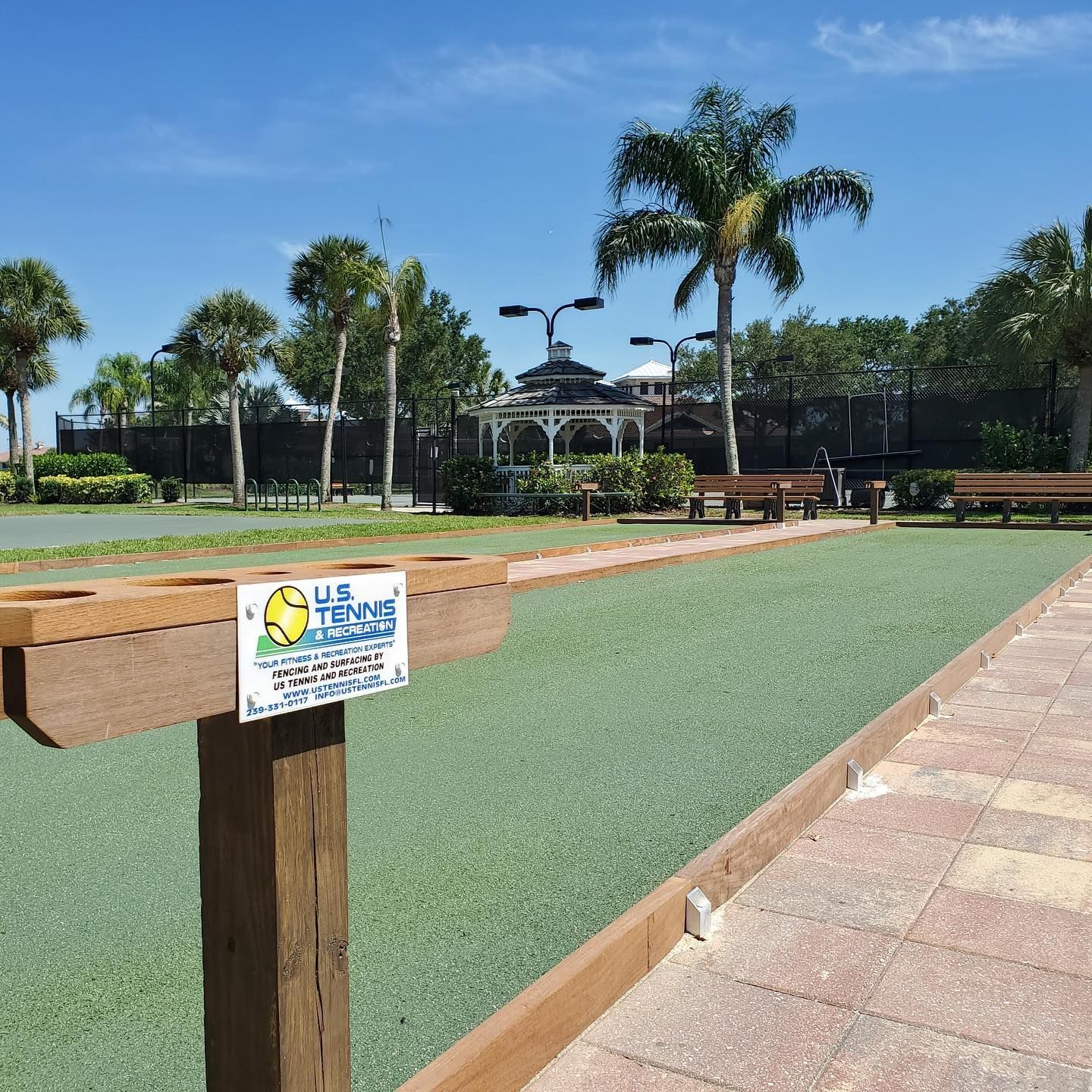 Sunny outdoor park with a mini-golf putting lane, palm trees, benches, and paved walkways.