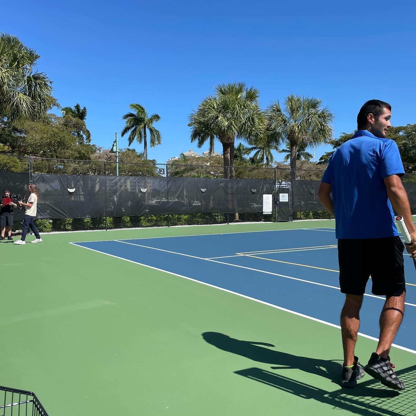 Outdoor tennis court with two players, one in blue in the foreground and one at the far side, under a clear sky