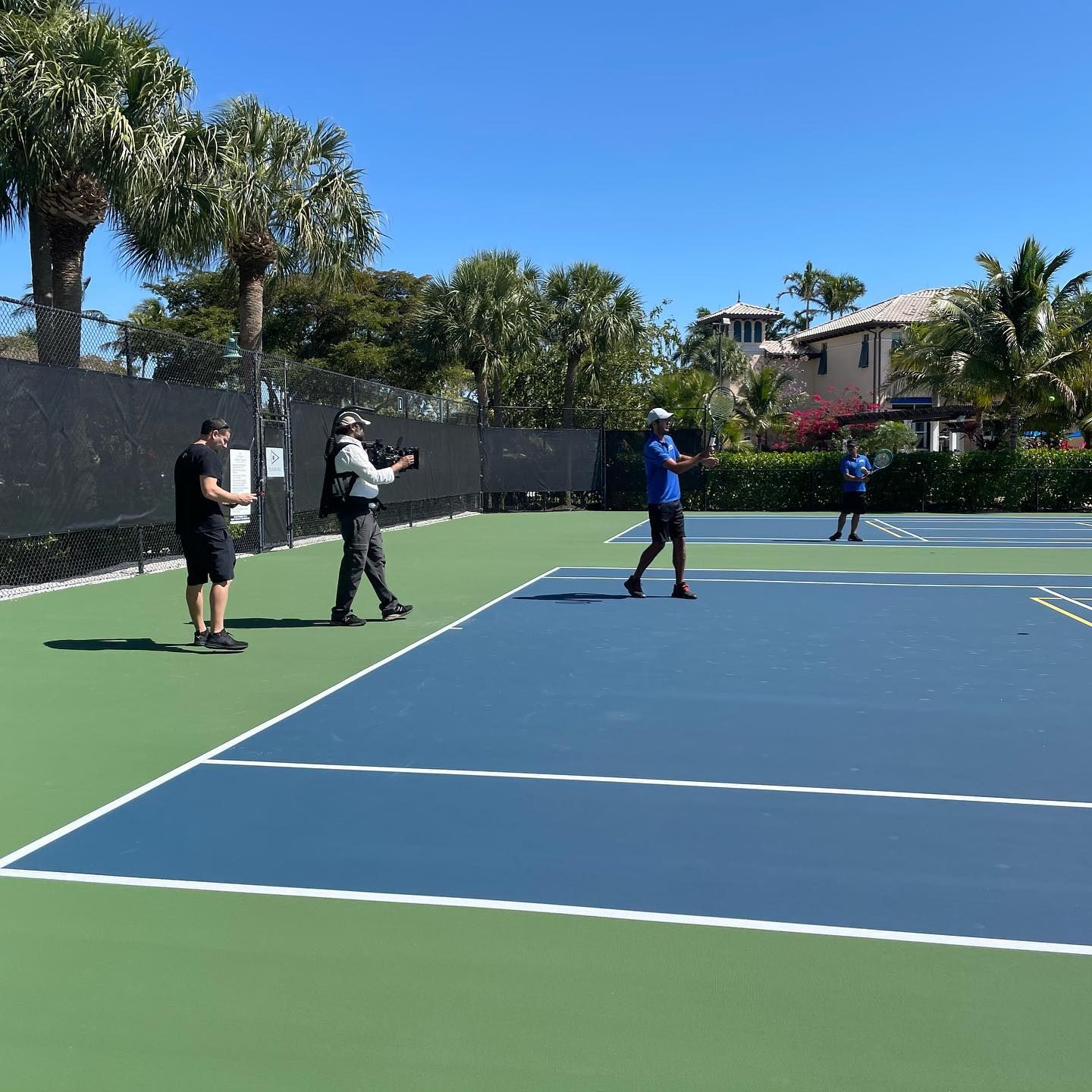 People standing on an outdoor tennis court under a clear blue sky, one holding a camera or phone.
