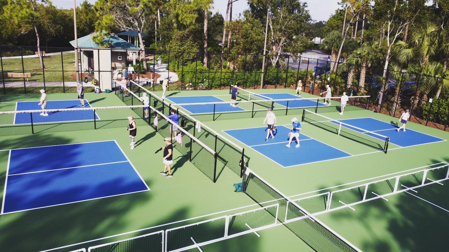 Outdoor pickleball courts with players on blue and green courts, surrounded by trees.
