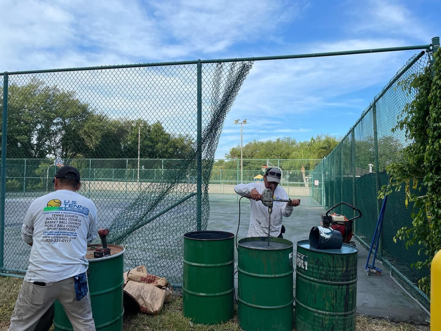 Two workers beside green barrels at an outdoor fenced court under a blue sky