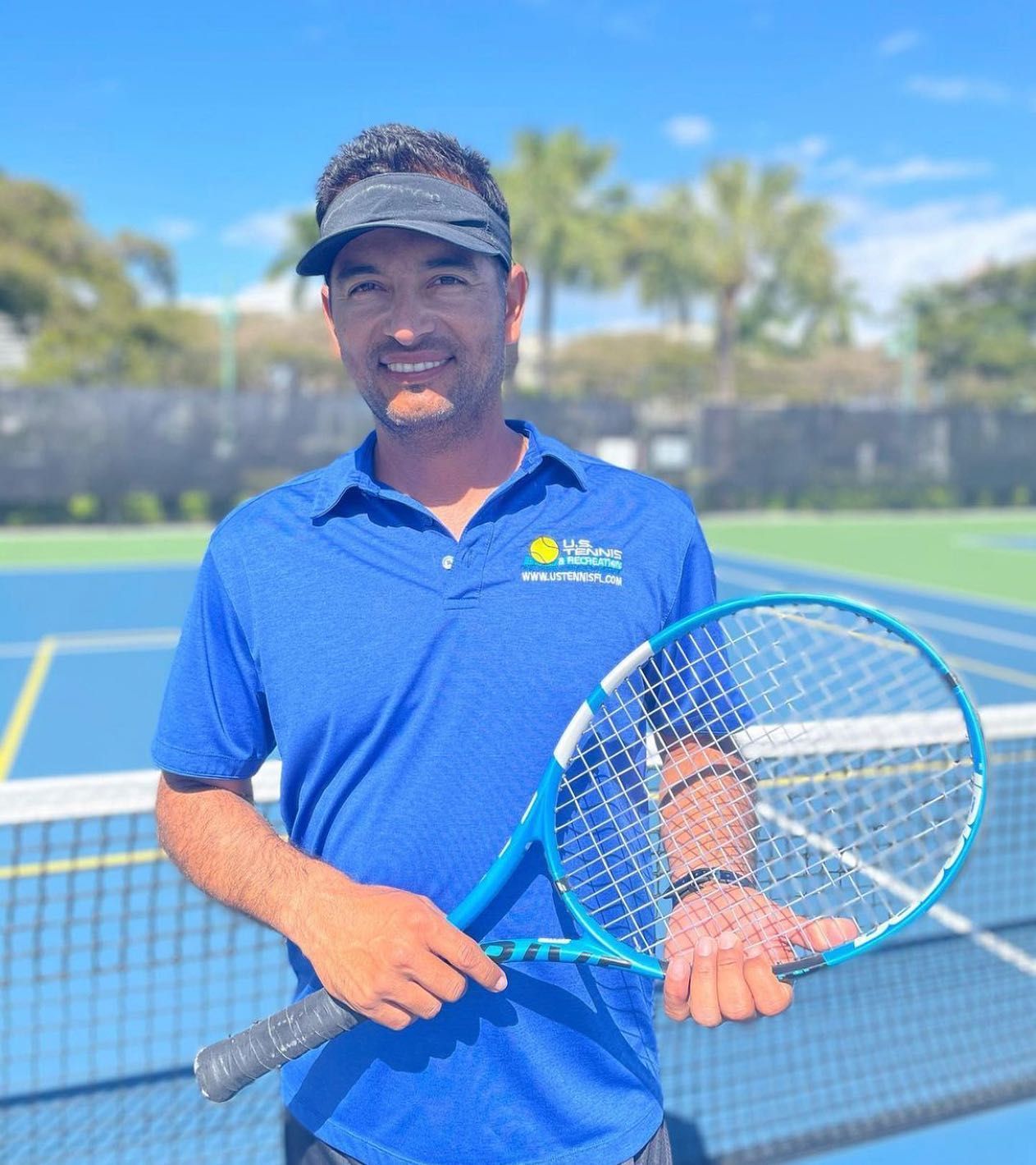 Man holding a tennis racket on an outdoor court, wearing a blue polo and cap.