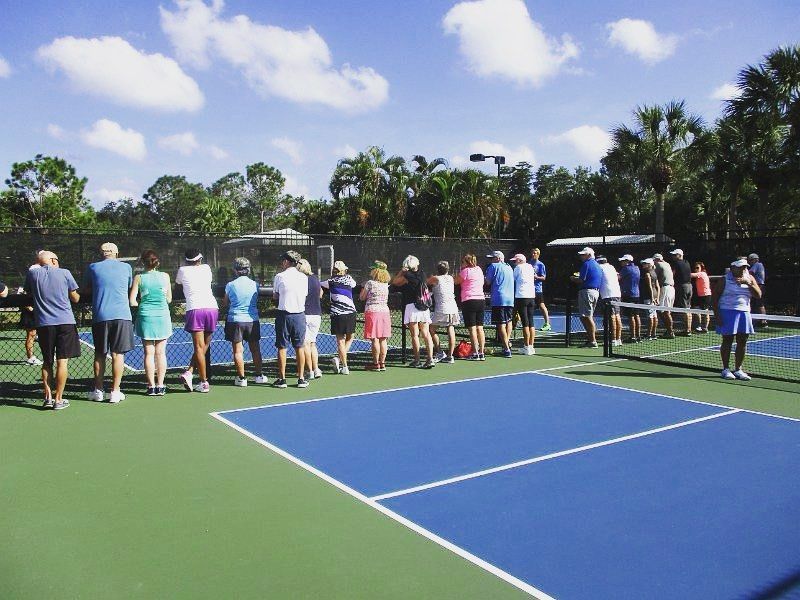 People standing along a tennis court fence under a bright blue sky, watching an outdoor event.