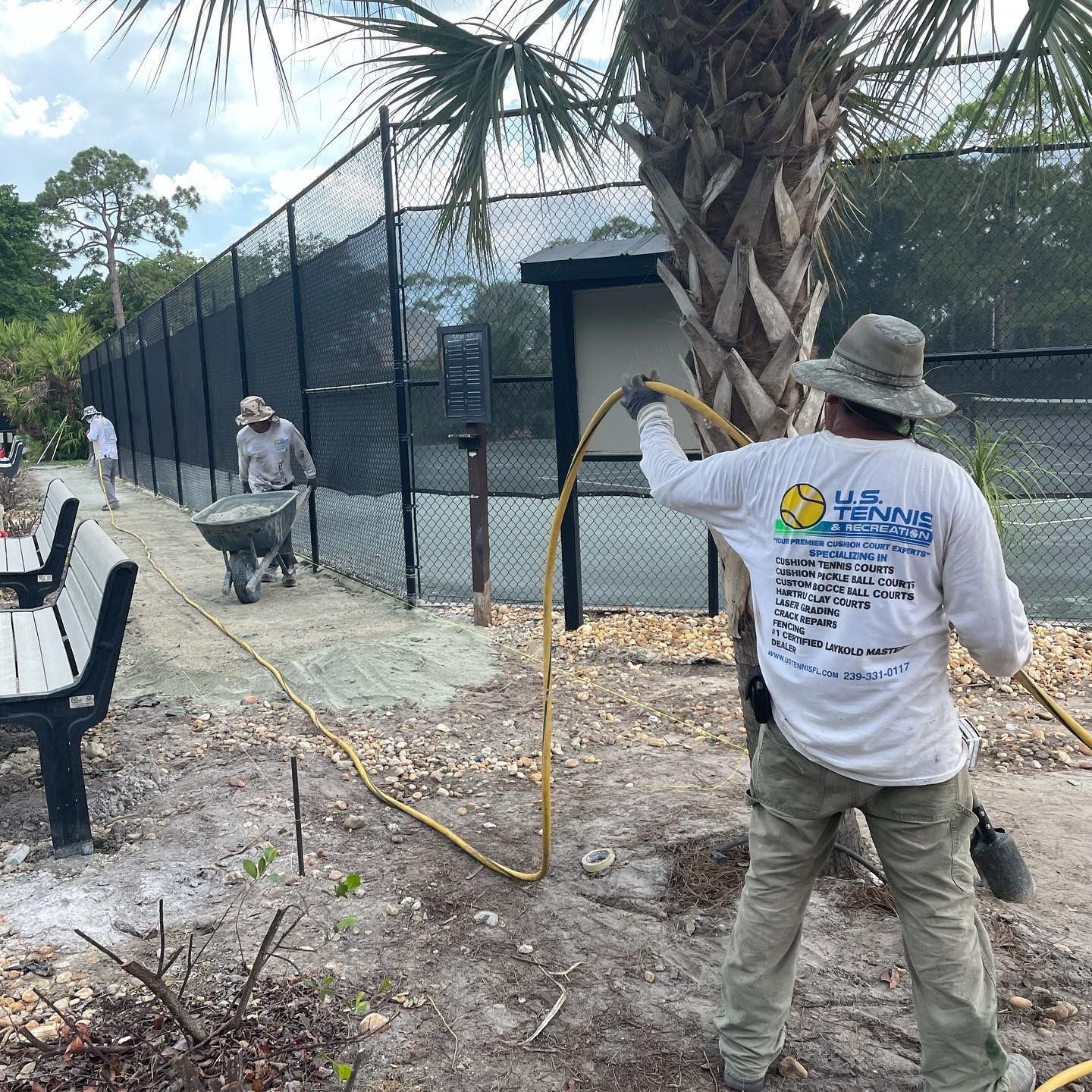 Workers using wheelbarrows and tools beside a black fence, spraying or laying material along a path.