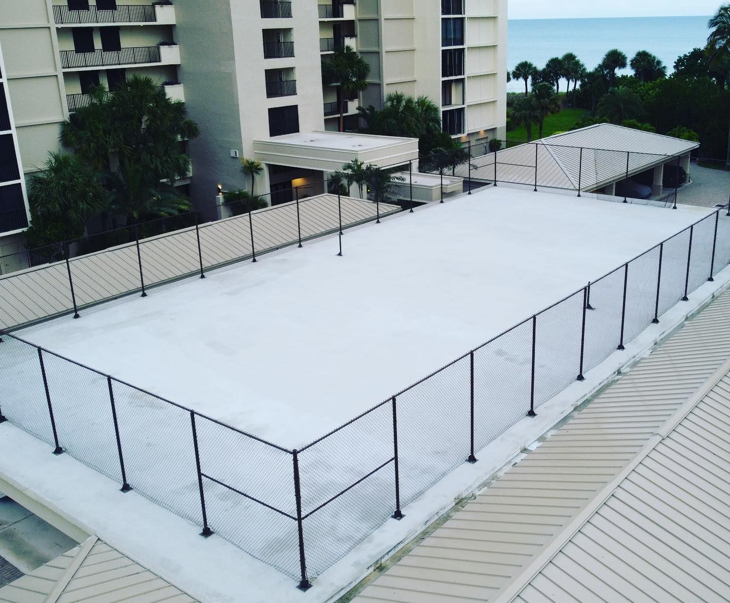 Empty fenced rooftop court with a white surface beside apartment buildings and trees