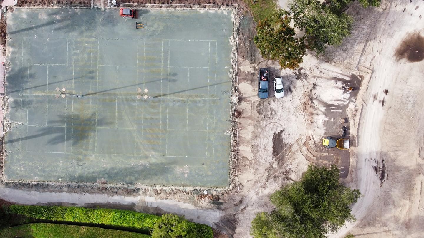 Aerial view of a worn outdoor tennis court beside trees, dirt paths, and a few parked vehicles